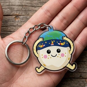 May include: A clear acrylic keychain featuring a cartoon character with a smiling face, pink cheeks, and a blue and green hat. The keychain has a silver metal ring and chain.