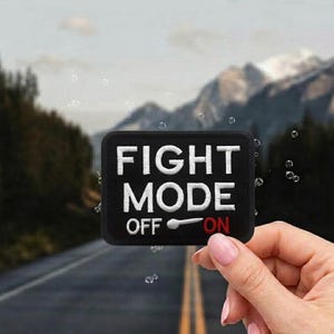 May include: Black rectangular patch with white embroidered text that reads "FIGHT MODE" with "OFF" and "ON" below. The switch is pointing to "ON". The background shows a road and mountains.