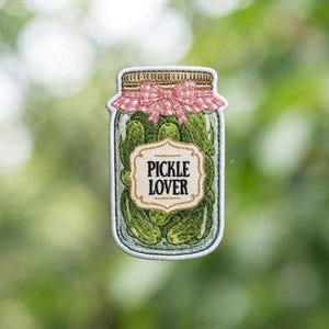 May include: Embroidered patch in the shape of a pickle jar, filled with green pickles. The jar has a pink and white gingham bow on top and the words "PICKLE LOVER" on a gold-bordered label.