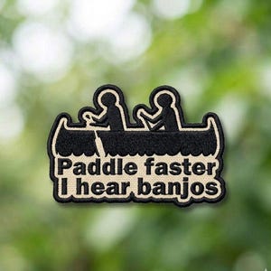 May include: Embroidered patch featuring a black canoe with two silhouetted figures paddling. The text "Paddle faster I hear banjos" is in a white font with a black border. The patch has a tan border.