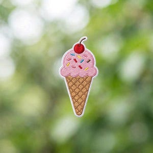 May include: Embroidered patch of a pink ice cream cone with sprinkles and a cherry on top. The cone is brown with a waffle pattern. The ice cream has colorful sprinkles. The patch has a white border.
