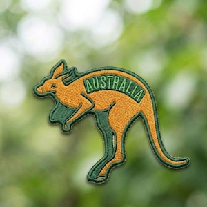 Puede incluir: Parche bordado con un canguro en naranja y verde. La palabra "AUSTRALIA" está arqueada sobre la espalda del canguro en letras verdes. El parche tiene una apariencia detallada y texturizada, adecuado para aplicar en ropa o accesorios.