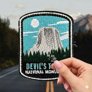 May include: Embroidered patch featuring Devil's Tower National Monument. The patch has a blue sky background with a white moon and clouds, a gray rock formation, and green trees. The text "DEVIL'S TOWER NATIONAL MONUMENT" is displayed in white.