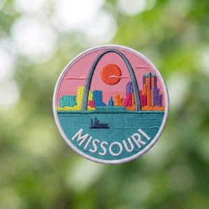 May include: A circular embroidered patch featuring a stylized St. Louis skyline with the Gateway Arch in grey, a bright orange sun, and colorful buildings against a pink sky. The lower portion depicts a teal river with a boat and the word MISSOURI.