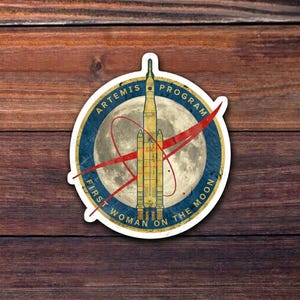May include: A sticker featuring the Artemis Program logo with a rocket, the moon, and the text "First Woman on the Moon." The design includes a blue and gold circular border with a red swoosh, set against a wooden background.