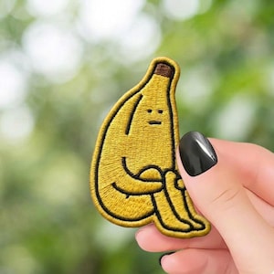 May include: Embroidered yellow banana patch with a sad face and arms wrapped around its knees. The patch has a brown stem and black outline. The patch is held by a person with black nail polish.