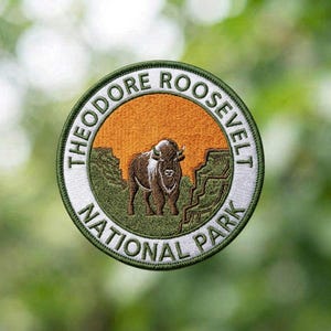 May include: A circular embroidered patch featuring a brown bison standing in a green landscape against an orange sky. The white border contains the black text THEODORE ROOSEVELT NATIONAL PARK, all enclosed within a dark green stitched edge.