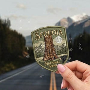 May include: A shield-shaped patch featuring a detailed illustration of a Sequoia tree, mountains, and a bear. The patch is embroidered with the words "SEQUOIA NATIONAL PARK" in a vintage style. The patch is held in front of a blurred background of a road and mountains.