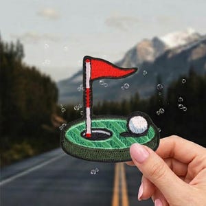 May include: Embroidered patch featuring a golf hole with a red flag, a white golf ball, and green grass. The patch is held up against a blurred background of a road and mountains.