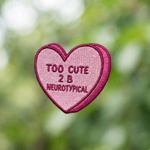 May include: A pink heart-shaped embroidered patch with a dark pink border. The patch features the text "TOO CUTE 2 B NEUROTYPICAL" in a darker pink thread. The background is a soft, blurred green.