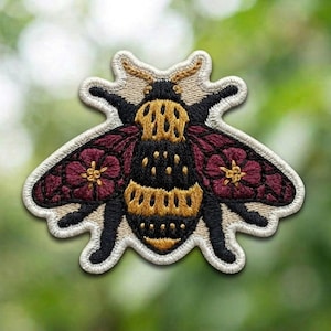May include: An embroidered patch shaped like a bee with a black and gold striped body. The wings are deep burgundy with floral patterns featuring yellow centers. The entire insect design is outlined in a cream-colored border with visible stitching.