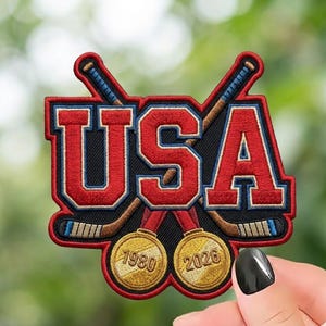 May include: Embroidered patch featuring the word "USA" in red, with crossed hockey sticks and two gold medals. The medals display the years 1980 and 2026. The patch has a black background and a red border.