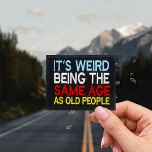 May include: A black square patch with the text "IT'S WEIRD BEING THE SAME AGE AS OLD PEOPLE" in white, red, and yellow. The patch is held in front of a blurred background of a road and mountains.