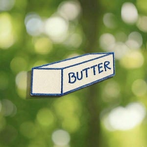May include: Embroidered patch in the shape of a butter stick. The patch is off-white with a blue border and the word "BUTTER" in blue lettering. The background is a blurred green.
