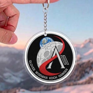 May include: A round keychain featuring a black and white embroidered design with a moon, Earth, and a red and white graphic. The keychain has the names "Glover Wiseman Koch Hansen" around the edge.