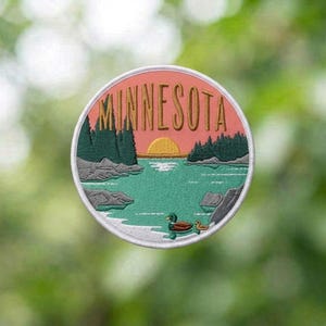 May include: A circular embroidered patch featuring a landscape scene with the word MINNESOTA in gold lettering at the top. The design includes a yellow sun, dark green pine trees, grey rocks, a teal lake, and two ducks swimming in the water.