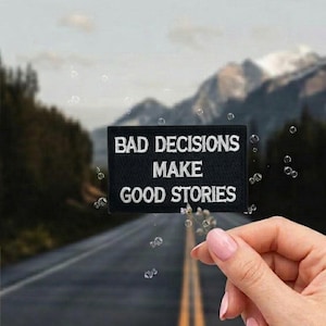May include: A black rectangular patch with white embroidered text that reads "BAD DECISIONS MAKE GOOD STORIES." The patch is held in front of a blurred background of a road, mountains, and sky.