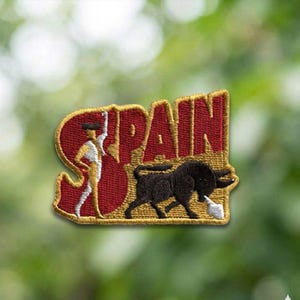 May include: Embroidered patch featuring the word "SPAIN" in red with a gold border. The patch depicts a matador and a black bull. The matador is wearing a red cape. The bull is charging with a white cloth.