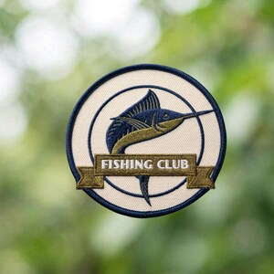 May include: Embroidered patch featuring a blue marlin leaping upwards. The design is set within a cream-colored circle, framed by a navy blue border. The words "FISHING CLUB" are displayed on a banner below the fish.