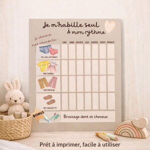 May include: A beige chart with the text "Je m'habille seul À mon rythme" and illustrations of clothing items. The chart includes spaces for each day of the week and the text "Prêt à imprimer, facile à utiliser". A plush bunny and wooden toys are in the foreground.