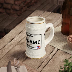 Puede incluir: Jarra de cerveza de cerámica blanca con asa, con un diseño que incluye el texto "Puerto Rico" y una placa de matrícula con la palabra "NAME" y la bandera de Puerto Rico. La jarra tiene ribetes dorados.