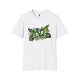 May include: White t-shirt featuring the word "UTUADO" in green, with tropical leaves and flowers. The text "Puerto Rico" is below the main text. The shirt has a crew neck and short sleeves.