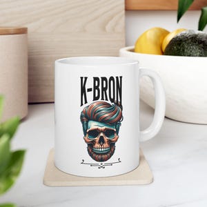 May include: White ceramic mug featuring a stylized skull with teal and orange hair and beard. The mug has the text "K-BRON" in black block letters above the skull design. The mug is on a beige coaster.