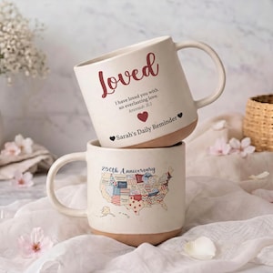 May include: Two stacked cream-colored ceramic mugs with speckled textures. The top mug features the word Loved in red script, the text I have loved you with an everlasting love. Jeremiah 31:3, a red heart, and Sarah's Daily Reminder. The bottom mug shows a map of the USA.