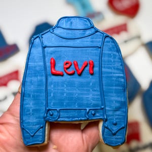 May include: A blue jacket-shaped cookie with the name "Levi" in red icing. The cookie is decorated with details that mimic a denim jacket. Other cookies are visible in the background, including heart-shaped cookies.