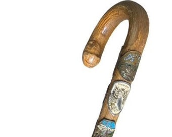 Puede incluir: Un bastón de madera con un mango curvo y varios pequeños distintivos metálicos unidos al eje. Los distintivos presentan varios diseños, incluyendo montañas, águilas y otros motivos inspirados en la naturaleza.
