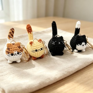 May include: Four round, crocheted cat keychains with metal rings. The cats are white with brown stripes, light yellow with a brown tail, solid black, and black with a white chest and tail tip. Each has small ears, black eyes, and stitched whiskers.