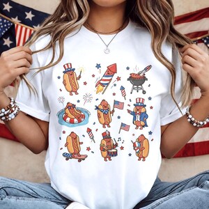 Pode incluir: Camiseta branca com um design divertido de cachorros-quentes de desenho animado celebrando o Dia da Independência. O design inclui cachorros-quentes com chapéus patrióticos, bandeiras e fogos de artifício. A camisa é um estilo casual e confortável.