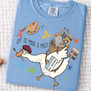 May include: A light blue t-shirt featuring a graphic of a white goose wearing a maroon beret and glasses, holding a paintbrush. The goose carries a basket of art supplies and a tag reading ART TEACHER. Text reads OFF TO MAKE A MESS.
