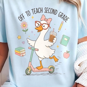 May include: A light blue t-shirt featuring a graphic of a white duck wearing red glasses and a pink bow, riding a scooter. The duck holds a coffee cup, surrounded by a flower, pencil, notebook, apple, and books with the text OFF TO TEACH SECOND GRADE.
