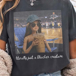 Puede incluir: Camiseta gris oscuro con un gráfico de una muñeca con gafas de sol y gorra de béisbol, sosteniendo un café helado. El texto "literally just a bleacher creature" está debajo. La camiseta es de estilo casual y unisex.