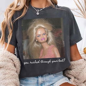 May include: Dark gray t-shirt featuring a distressed image of a doll with a displeased expression. The text "you rushed through your test?" is below the image. The shirt is worn with denim shorts.