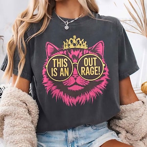 May include: Dark gray t-shirt featuring a pink cat face wearing sunglasses and a gold crown. The sunglasses have the text "THIS IS AN OUT RAGE!". The shirt is a casual style.