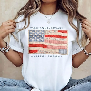 May include: A white short-sleeve t-shirt featuring a graphic of an American flag composed of various patterned fabric patches. The text 250TH ANNIVERSARY is printed above the flag, and the dates 1776 - 2026 are printed below in dark blue lettering.