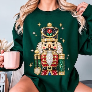 May include: A dark green long-sleeve sweatshirt featuring a central graphic of a nutcracker adorned with sparkling, multi-colored rhinestones. The nutcracker design includes a red, gold, and black uniform with a white beard and star accents.