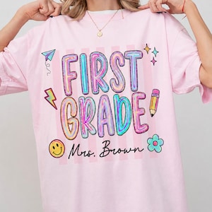 May include: A light pink t-shirt featuring the text FIRST GRADE in large, colourful, bubble-style letters. Below the text, the name Mrs. Brown is written in black script. The design includes a paper aeroplane, lightning bolt, pencil, and smiley face.