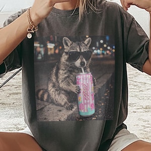 Puede incluir: Camiseta gris oscuro con un estampado de un mapache con gafas de sol bebiendo de una lata. La lata tiene un diseño rosa y azul con la palabra "Always". El mapache está sentado en una superficie de piedra.