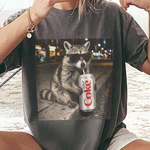 Op de afbeelding: Donkergrijze t-shirt met een afbeelding van een wasbeer met een zonnebril, drinkend uit een Diet Coke-blikje. De wasbeer staat tegen een vage stadsachtergrond. Het shirt heeft een relaxte, oversized pasvorm.