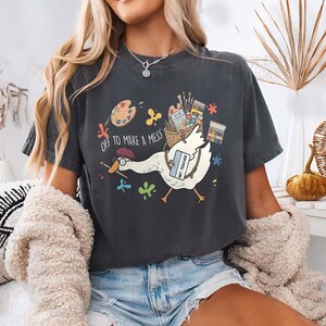 May include: A charcoal gray t-shirt featuring a graphic of a white goose wearing a red beret and glasses, carrying a basket of art supplies. The text reads "OFF TO MAKE A MESS" and "ART TEACHER" on a name tag, surrounded by colorful paint splatters.