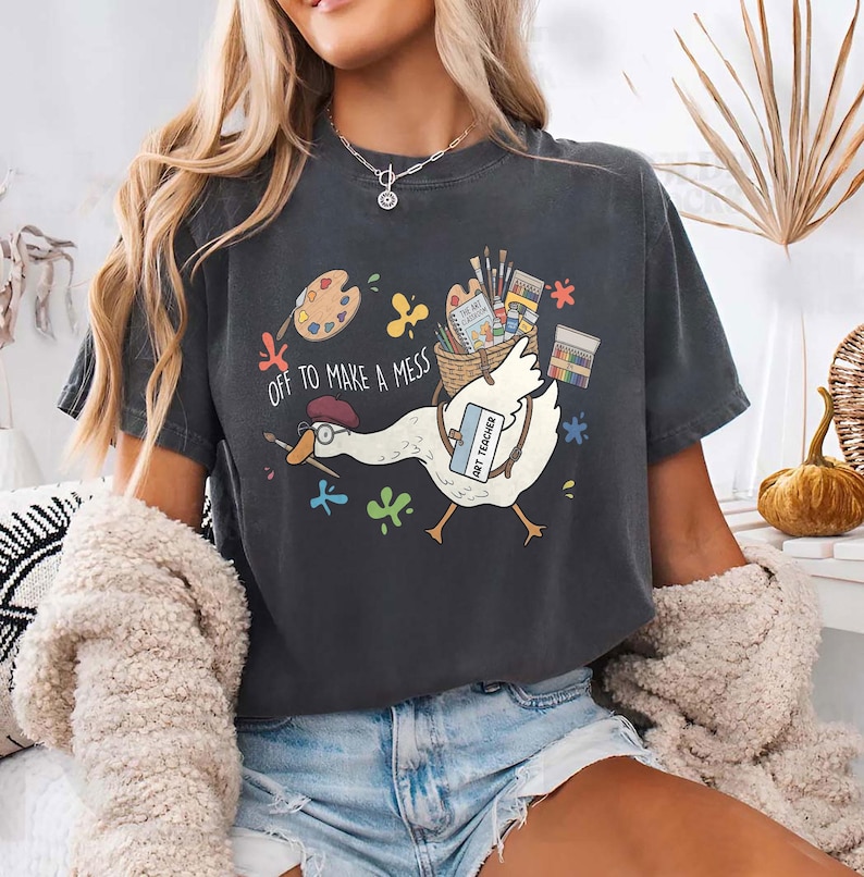 May include: A charcoal gray t-shirt featuring a graphic of a white goose wearing a red beret and glasses, carrying a basket of art supplies. The text reads "OFF TO MAKE A MESS" and "ART TEACHER" on a name tag, surrounded by colorful paint splatters.