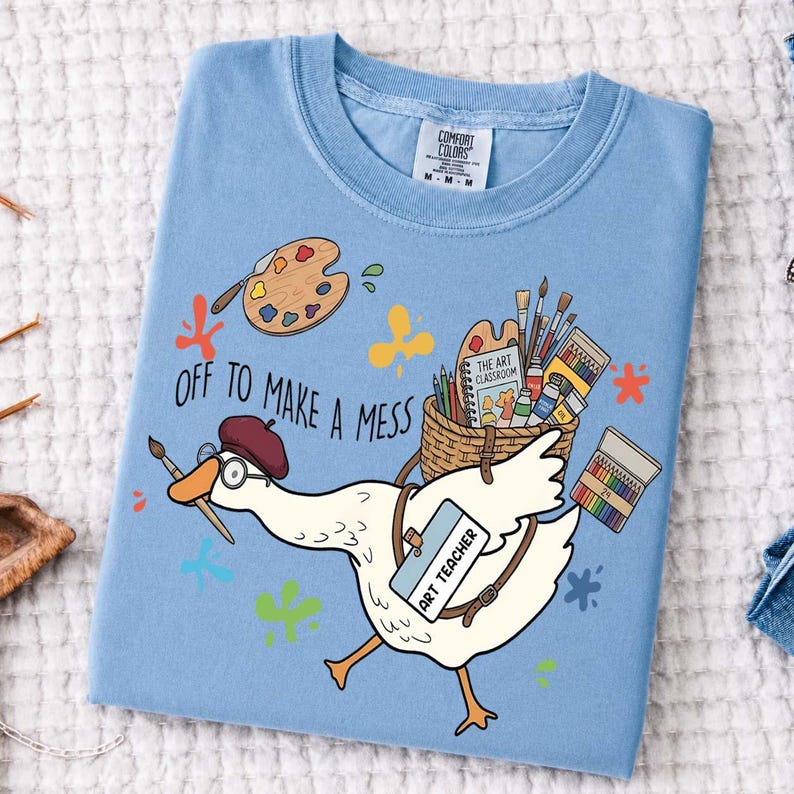 May include: A light blue t-shirt featuring a graphic of a white goose wearing a maroon beret and glasses, holding a paintbrush. The goose carries a basket of art supplies and a tag reading ART TEACHER. Text reads OFF TO MAKE A MESS.