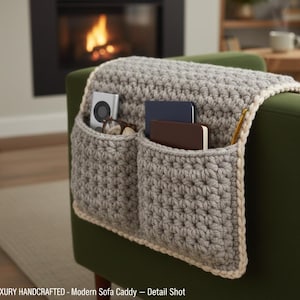 Può includere: Un organizer per divano grigio lavorato a mano all'uncinetto con due tasche, appoggiato su un divano verde. L'organizer ha un bordo color crema e contiene un piccolo lettore multimediale, occhiali e quaderni. È visibile un uncinetto.