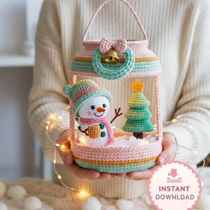 May include: A crocheted Christmas lantern in pastel colors, featuring a snowman and Christmas tree inside. The lantern has a handle, a bow, and a small bell. The text "Christmas Lantern - CROCHET PATTERN" is displayed at the bottom.
