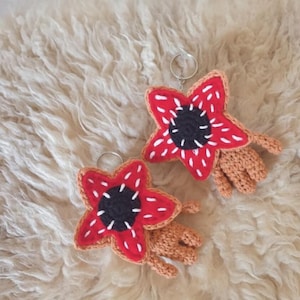 May include: Two handmade Demogorgon keyrings. Each keyring features a red, star-shaped flower with white accents and a black centre. The keyrings have tan-coloured, crocheted hands. The keyrings are on a fluffy, cream-coloured surface.