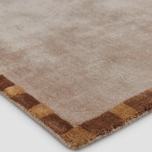 May include: A corner view of a beige rug with a brown and tan checkered border. The rug has a soft, plush texture and a neutral color palette, suitable for various interior design styles.