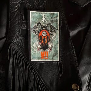May include: A rectangular patch with a motorcycle graphic in orange, black, and white, with the text "RUST NOIRE" and "MII" on a teal background. The patch is affixed to a black leather jacket with fringe.